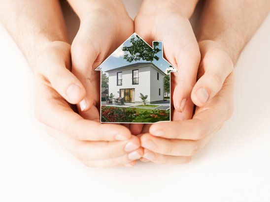 Symbolisches Bild: ein Miniaturhaus in schützende Hände - Sinnbild für Sicherheit und Geborgenheit beim Hausbau.